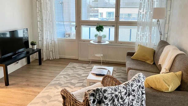 A modern living room with a flat-screen TV, a sofa with cushions, a coffee table, and a window with a view of buildings. - Kotimaailma KotikaksioKirkkok saunalla (Lahti)
