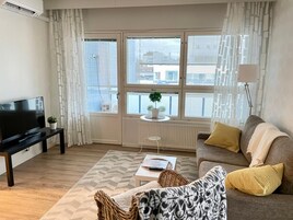 Moderni olohuone, jossa on litteänäyttöinen televisio, tyynyillä varustettu sohva, sohvapöytä ja ikkuna, josta on näkymä rakennuksiin.