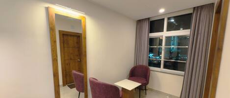 A hotel room with a wooden door, a large mirror, two purple chairs, a small table, and a window with curtains.