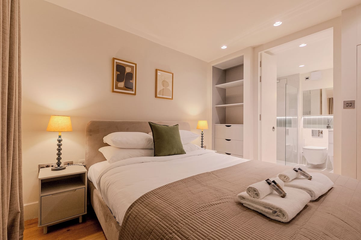 Cama bem arrumada com um espalha cobre marrom, duas luminárias de cabeceira, um criado-mudo e um banheiro visível através de uma porta aberta.