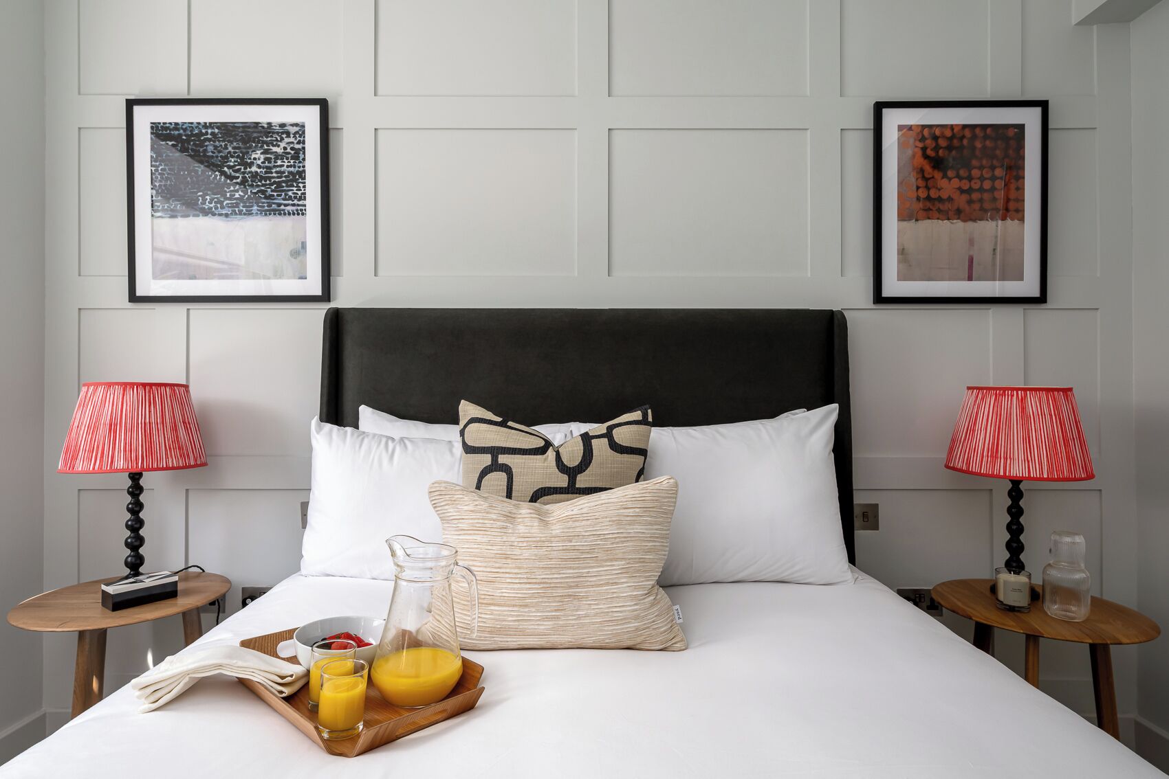 A neatly made bed with a black headboard, two red lamps, and a tray with breakfast items.