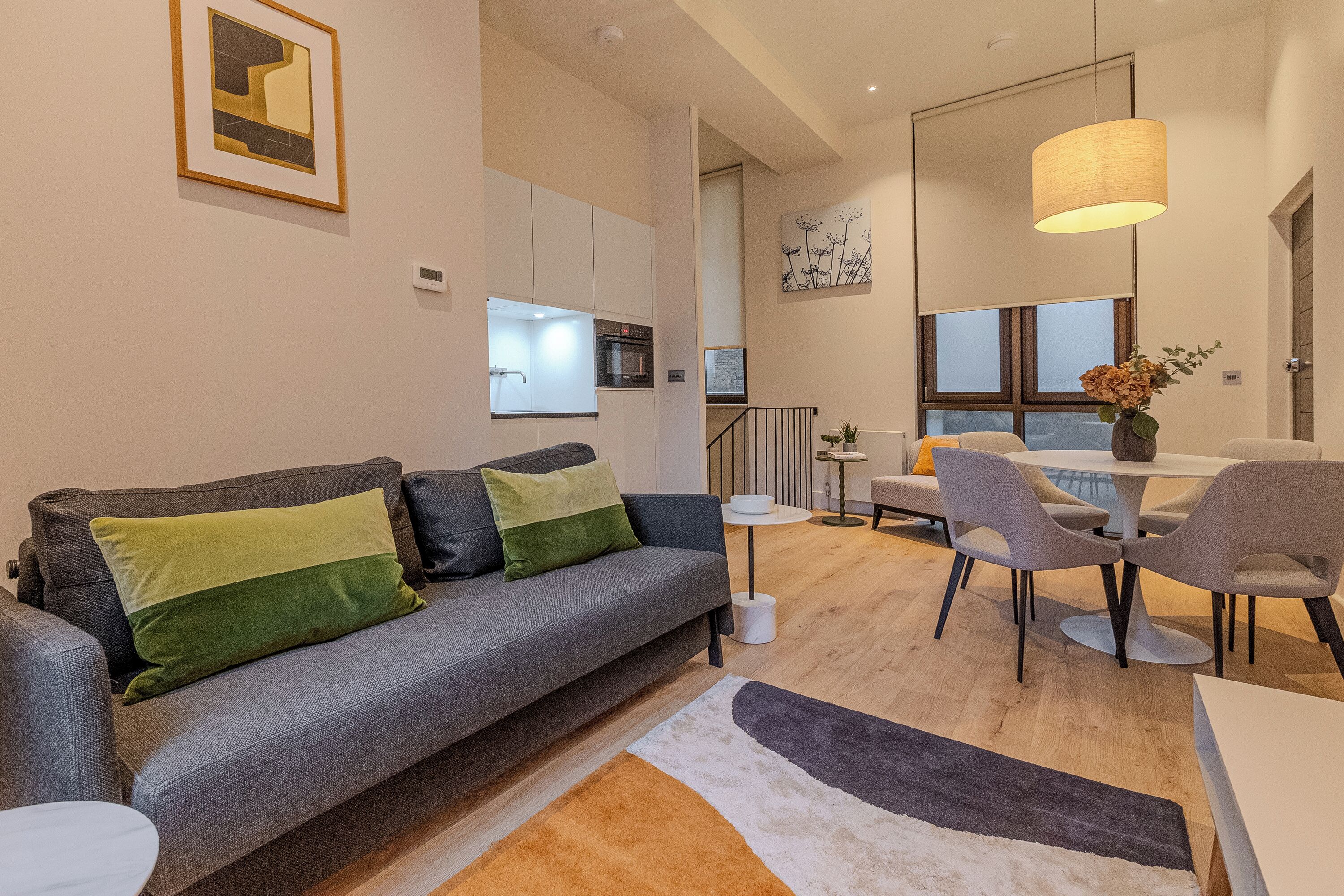 A modern living room with a grey sofa, a dining table with chairs, a kitchen in the background, and a large window.