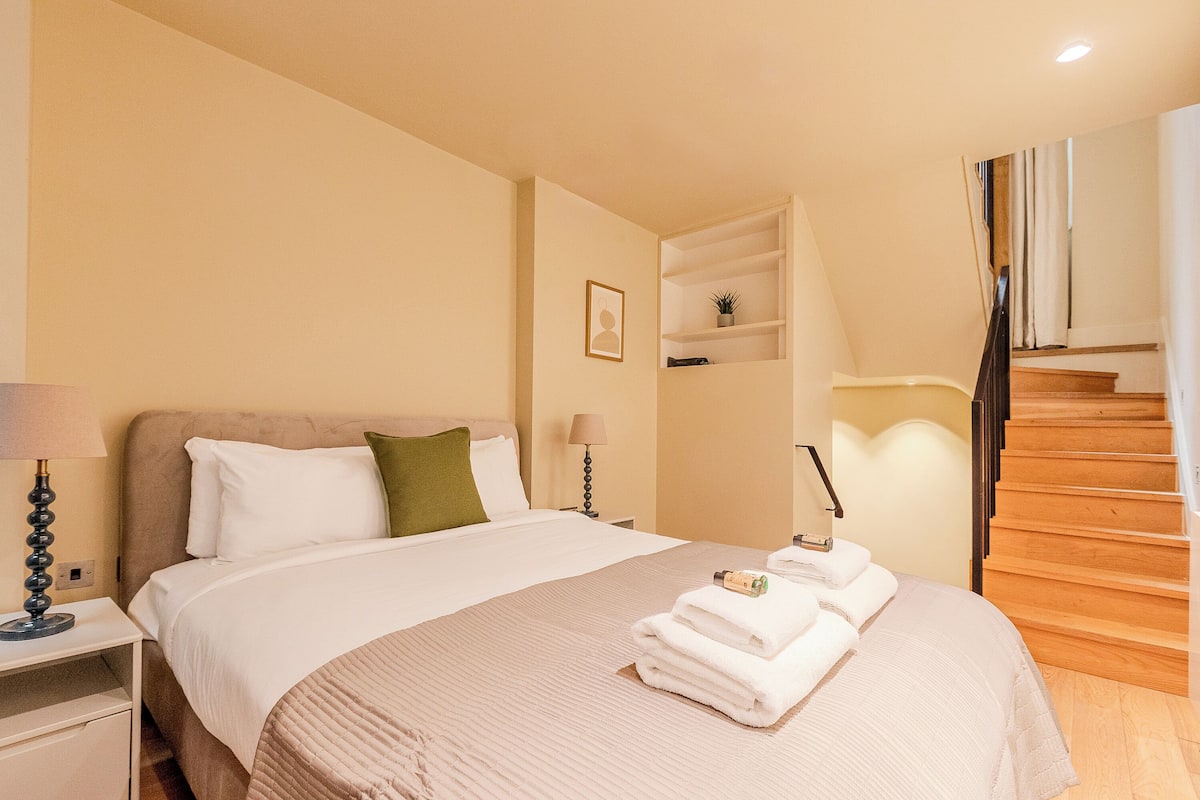Quarto de hotel com cama, mesas de cabeceira, abajur, prateleira pequena com planta e escada.