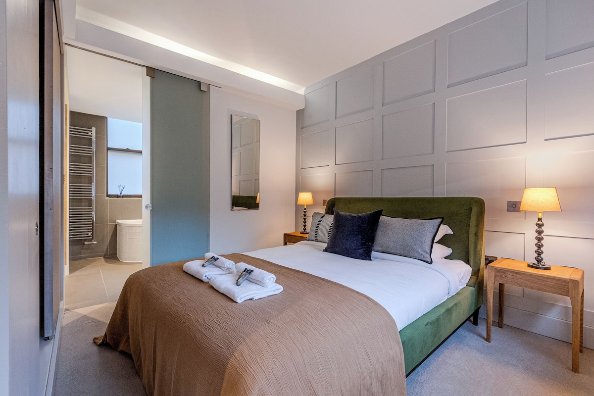 Quarto de hotel moderno com uma cama grande, duas mesas de cabeceira com abajures, um espelho e vista para um banheiro.