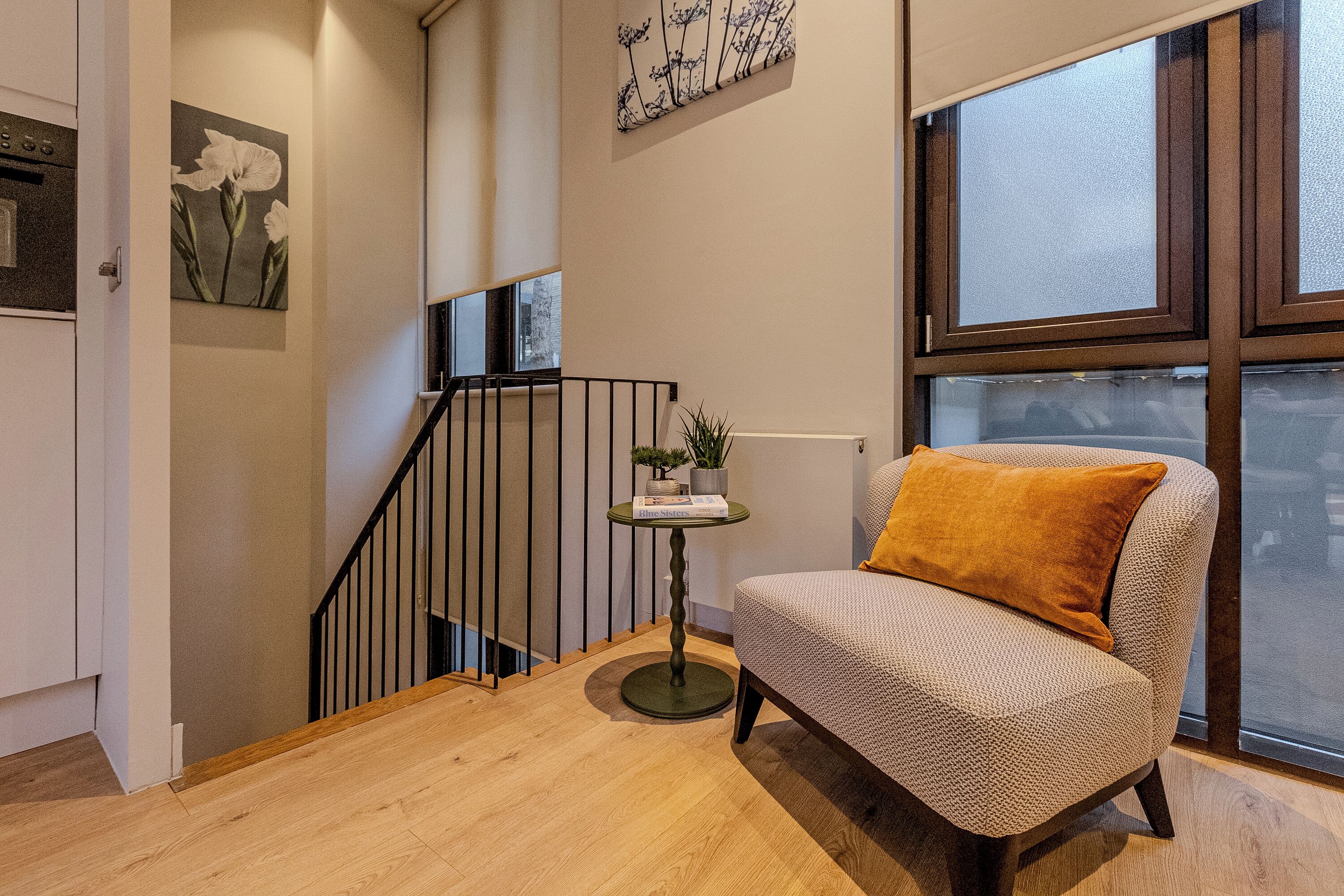 Un salón moderno con piso de madera, un sillón estampado con cojín, una mesita con una planta y una escalera con barandilla.