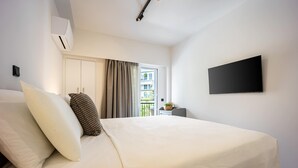 A hotel room with a large bed, a flat-screen TV mounted on the wall, a window with curtains, and a small table with a plant. - Stylish 1BR in Pangrati | Balcony & Green View (Athina)