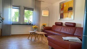 Ein modernes Wohnzimmer mit einem Ledersofa, einem kleinen Holztisch mit einer Vase voller Sonnenblumen und einem großen Fenster mit Blick auf Bäume.
