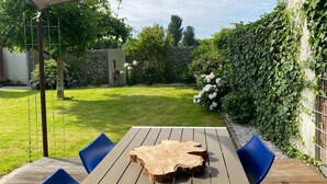 Ein Holztisch mit einem besonderen Mittelstück, umgeben von blauen Stühlen, unter einem schattigen Bereich mit Blick auf einen gepflegten Garten und eine Steinmauer.