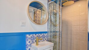 Ein Badezimmer mit einem weißen Waschbecken, blauen und weißen Fliesen mit Muster sowie einem runden Spiegel und einer Duschkabine aus Glas.