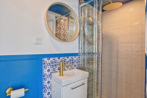 A bathroom with a white sink, blue and white patterned tiles, a round mirror, and a glass shower enclosure. - Bleu Horizon - Studio with balcony and sea view (Villers-sur-Mer)