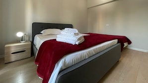 Una cama bien tendida con un edredón rojo, una mesita de noche con una lámpara y un aplique de pared.