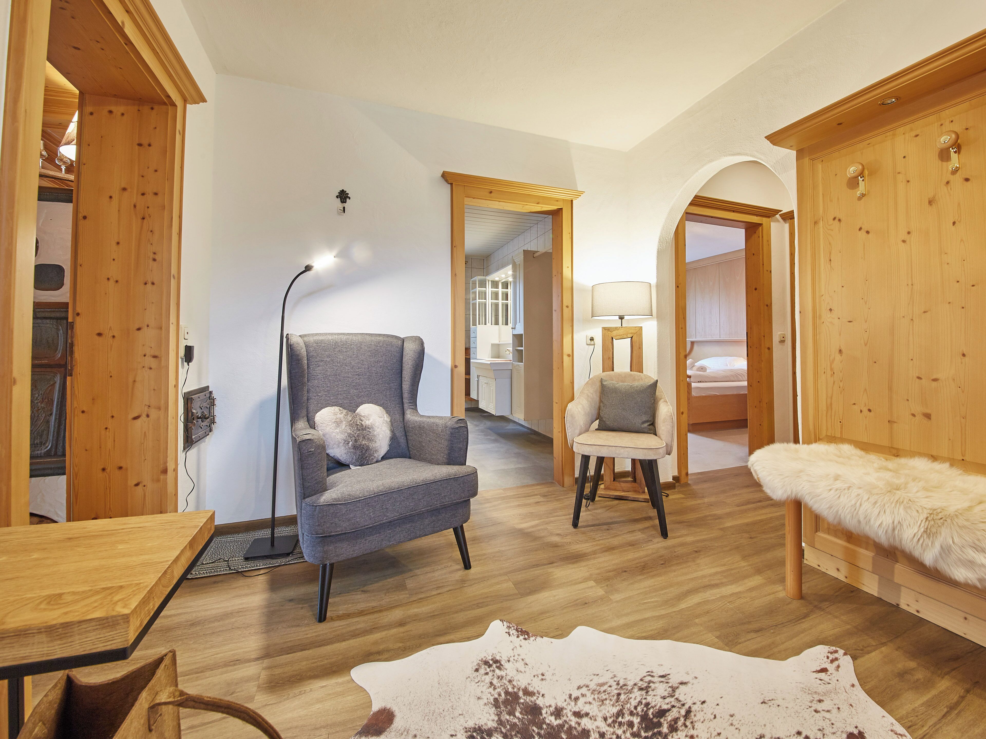 A room with a wooden floor, a grey armchair with a heart-shaped pillow, a white armchair, a wooden wardrobe, and a fur-covered bench.