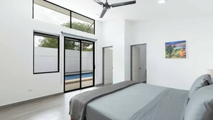 A modern bedroom with a large bed, a ceiling fan, and a view of a swimming pool through glass doors. - Casa Aurora with Private Patio & Pool (Playas Del Coco)