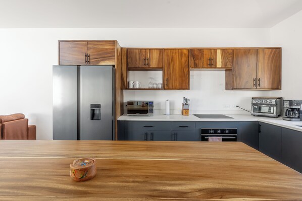 A modern kitchen with wooden cabinets, a stainless steel refrigerator, a microwave, a toaster, and a wooden dining table.