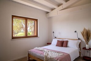 Quarto com cama de madeira, janela com vista para árvores, mesa de cabeceira com abajur e um vaso com plumas de capim-do-campo.
