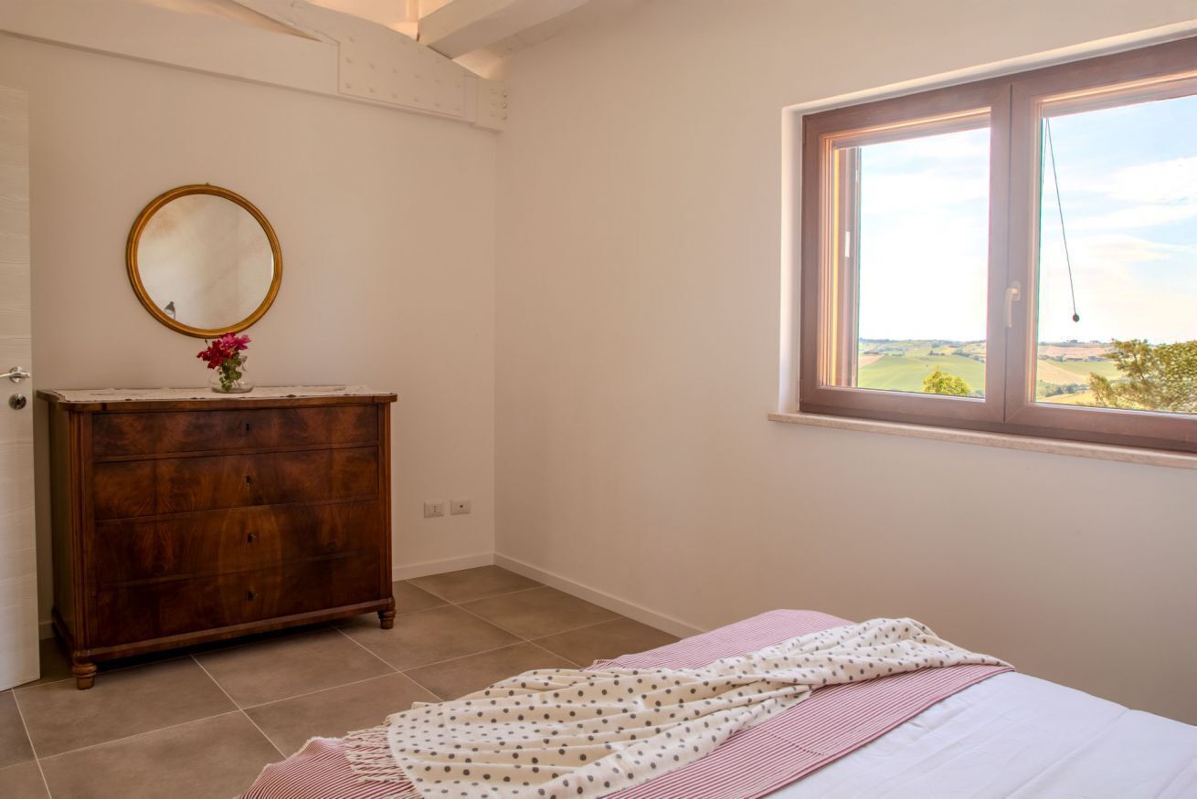 Une chambre avec une commode en bois, un miroir rond, un lit recouvert d’une couverture à pois et une fenêtre donnant sur la campagne.