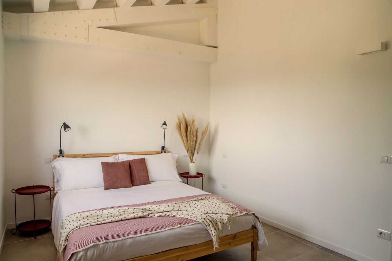Un dormitorio con una cama de madera, dos mesitas de noche, una lámpara roja y un florero con plantas secas.