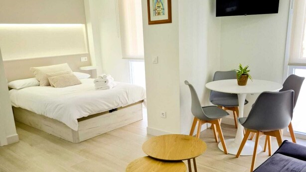 A modern hotel room with a bed, a small dining table, two chairs, a sofa, and a TV mounted on the wall.