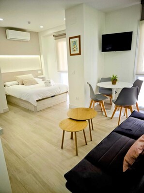 Una habitación de hotel moderna con una cama, una pequeña mesa de comedor, dos sillas, un sofá y un televisor colgado en la pared.