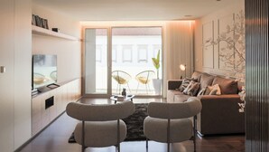Ein modernes Wohnzimmer mit einer Couch, zwei Sesseln, einem Couchtisch und einem Fernseher. Ein großes Fenster bietet Blick auf die umliegenden Gebäude.