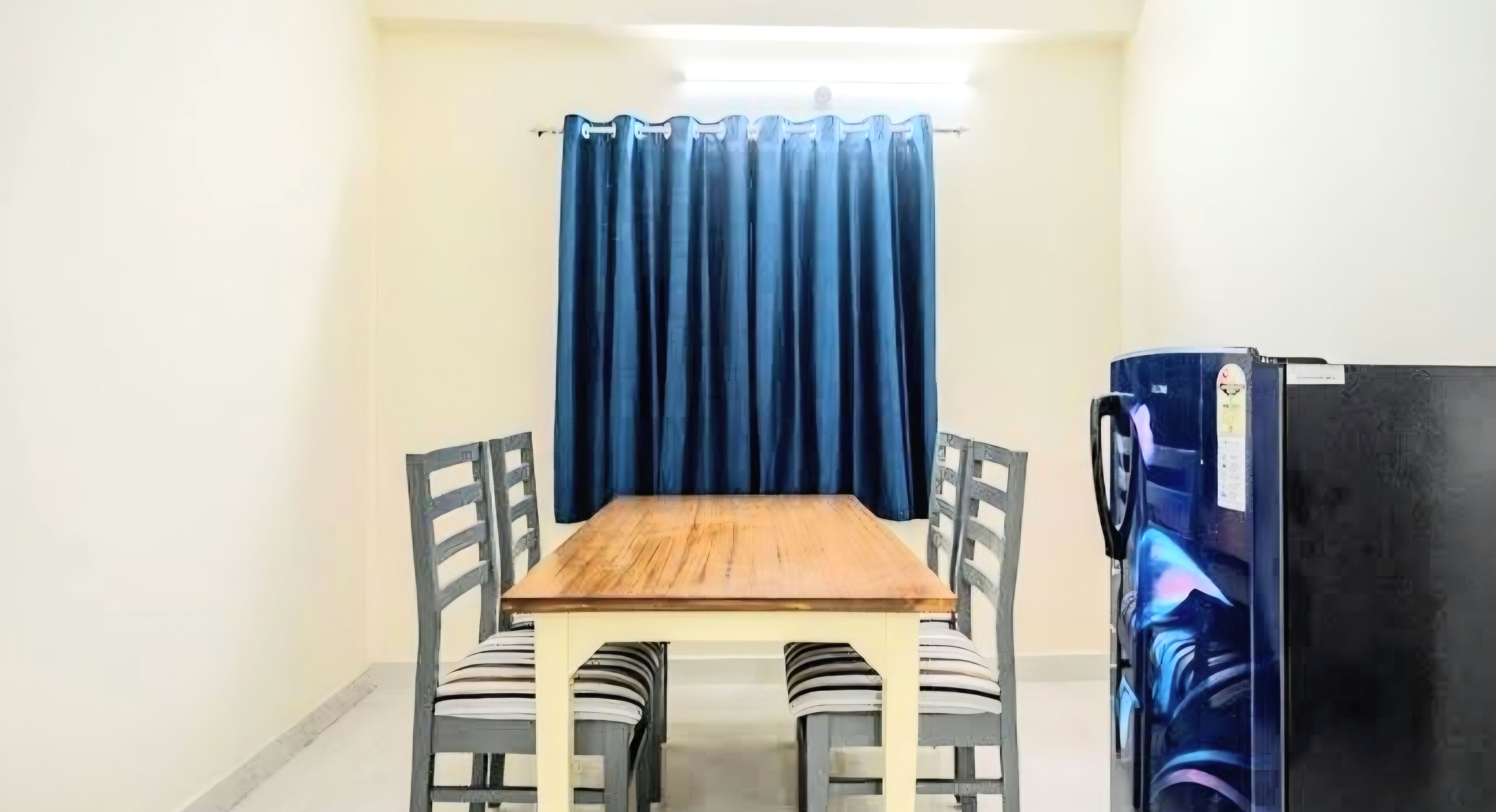 Una habitación pequeña con una mesa de madera y cuatro sillas, un cortin azul y un refrigerador.