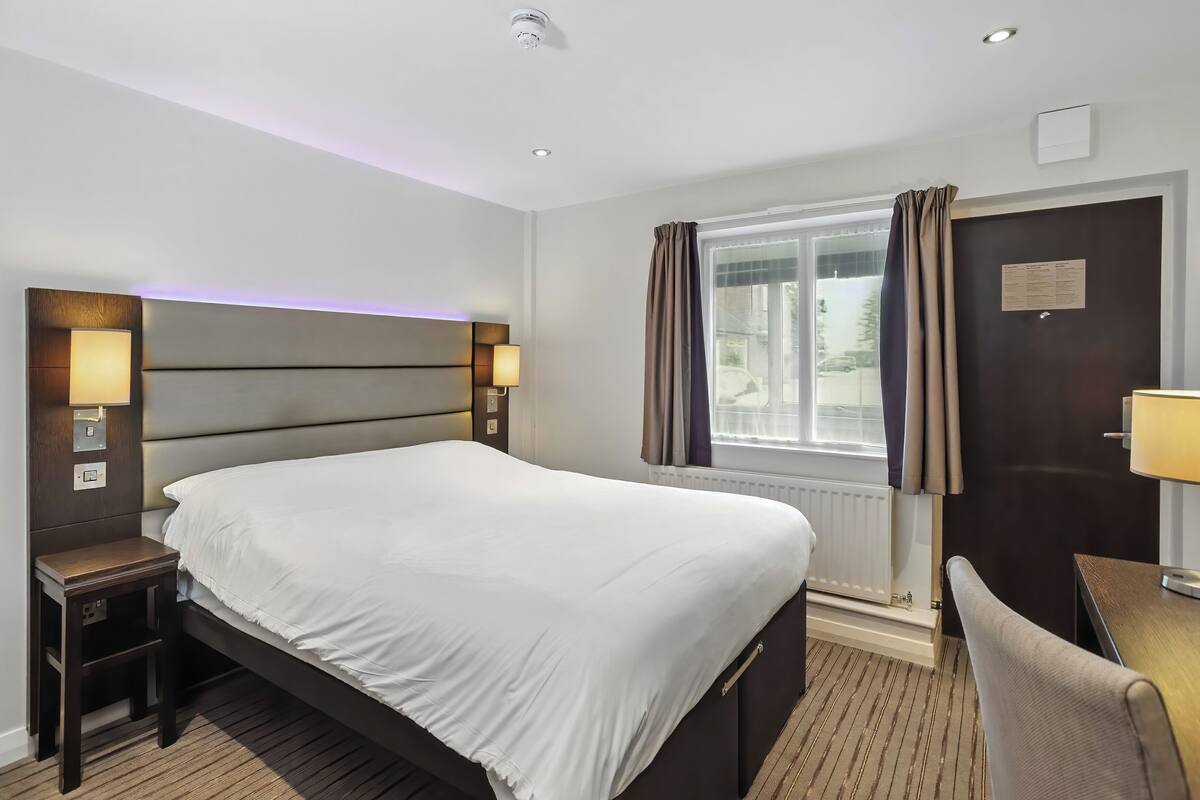 Ein Hotelzimmer mit einem großen Bett, Nachttischen mit Lampen, einem Schreibtisch und einem Fenster mit Vorhängen.
