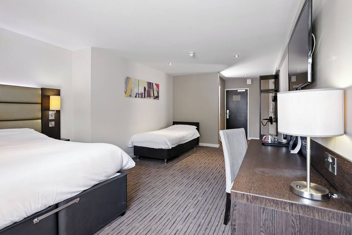 Ein Hotelzimmer mit zwei Betten, einem Flachbildfernseher, einem Schreibtisch mit Lampe und einem Bild an der Wand.