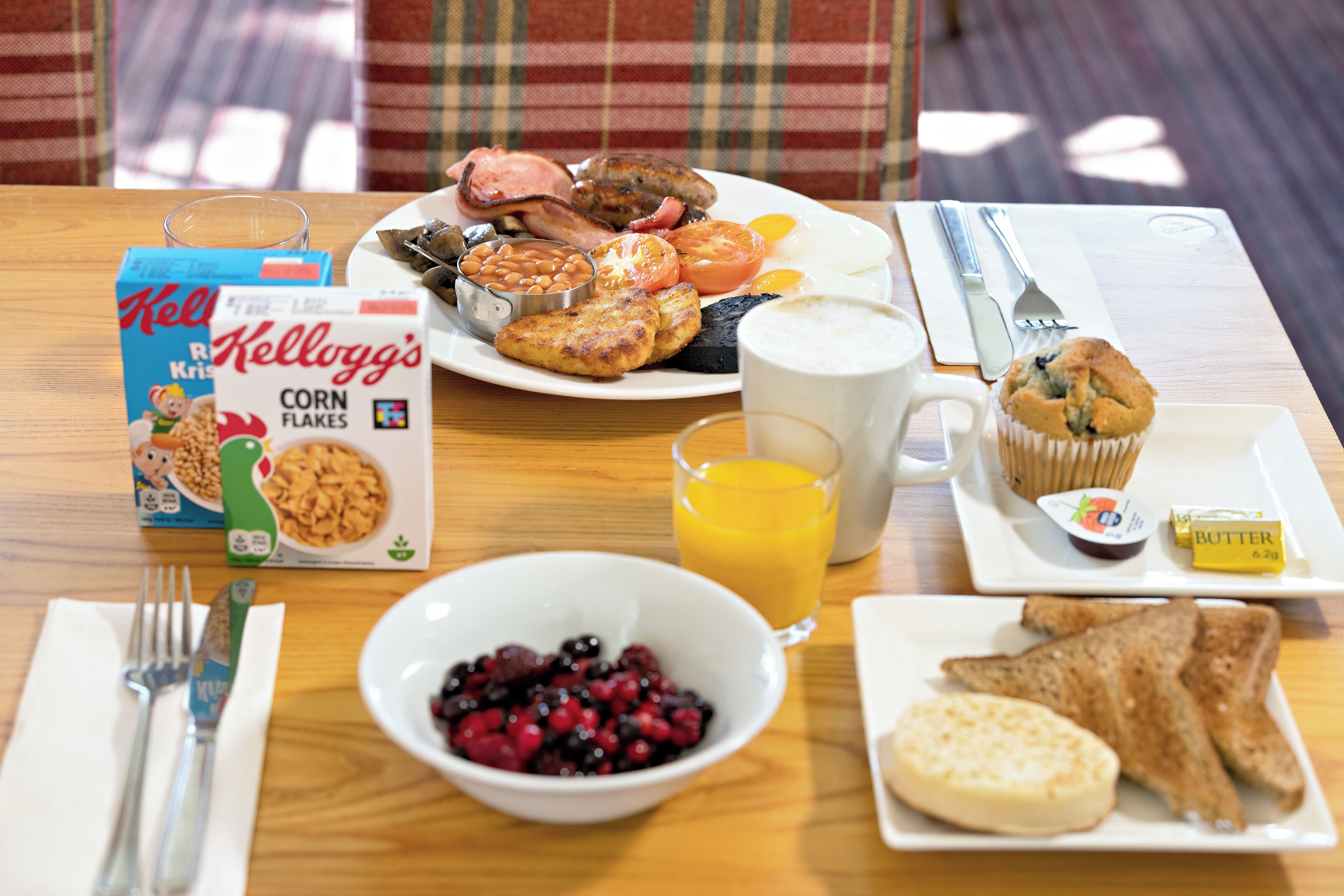 A breakfast table with a box of Kellogg's corn flakes, a bowl of berries, a glass of orange juice, toast, a muffin, and a plate with eggs, beans, and tomatoes.
