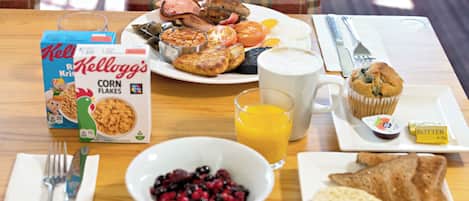 A breakfast table with a box of Kellogg's corn flakes, a bowl of berries, a glass of orange juice, toast, a muffin, and a plate with eggs, beans, and tomatoes.