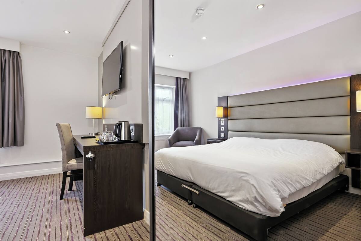 Ein Hotelzimmer mit einem großen Bett, einem Schreibtisch mit Stuhl, einem an der Wand montierten Fernseher und einem Fenster mit Vorhängen.