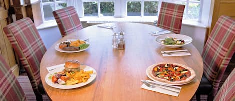 Una mesa redonda de madera con cuatro sillas, cada una con tapicería escocesa. Hay platos de comida, incluyendo una hamburguesa, papas fritas y una pizza, además de salsas y cubiertos.