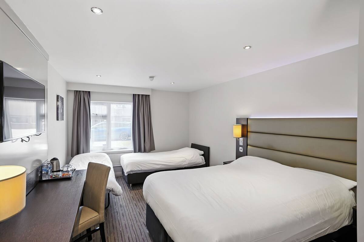Ein Hotelzimmer mit zwei Betten, einem Schreibtisch mit Lampe, einem Sessel, einem Fenster mit Vorhängen und einem an der Wand befestigten Fernseher.
