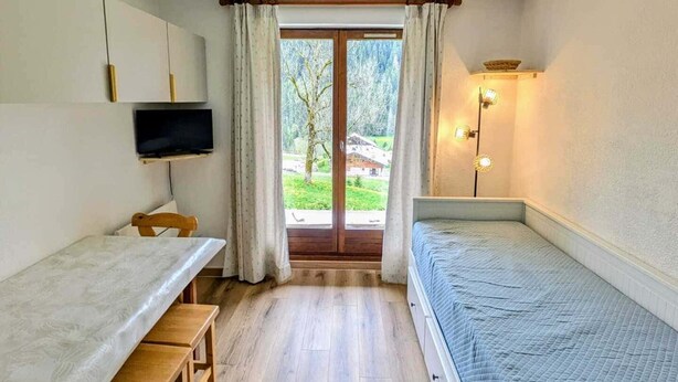 A small room with a bed, a desk, a chair, a television, and a window with curtains. - Cozy studio for 4 in Châtel with terrace and parking (Châtel)
