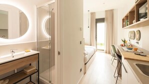 Ein modernes Hotelzimmer mit einem Badezimmer, das eine gläserne Dusche, einen großen Spiegel und einen Waschtisch mit Waschbecken bietet. Das Zimmer verfügt über ein Bett, einen Schreibtisch mit Stuhl und Blick ins Freie.