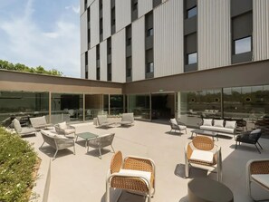 Une terrasse extérieure moderne, dotée de meubles en osier, de murs en verre et d’un bâtiment à la façade quadrillée.