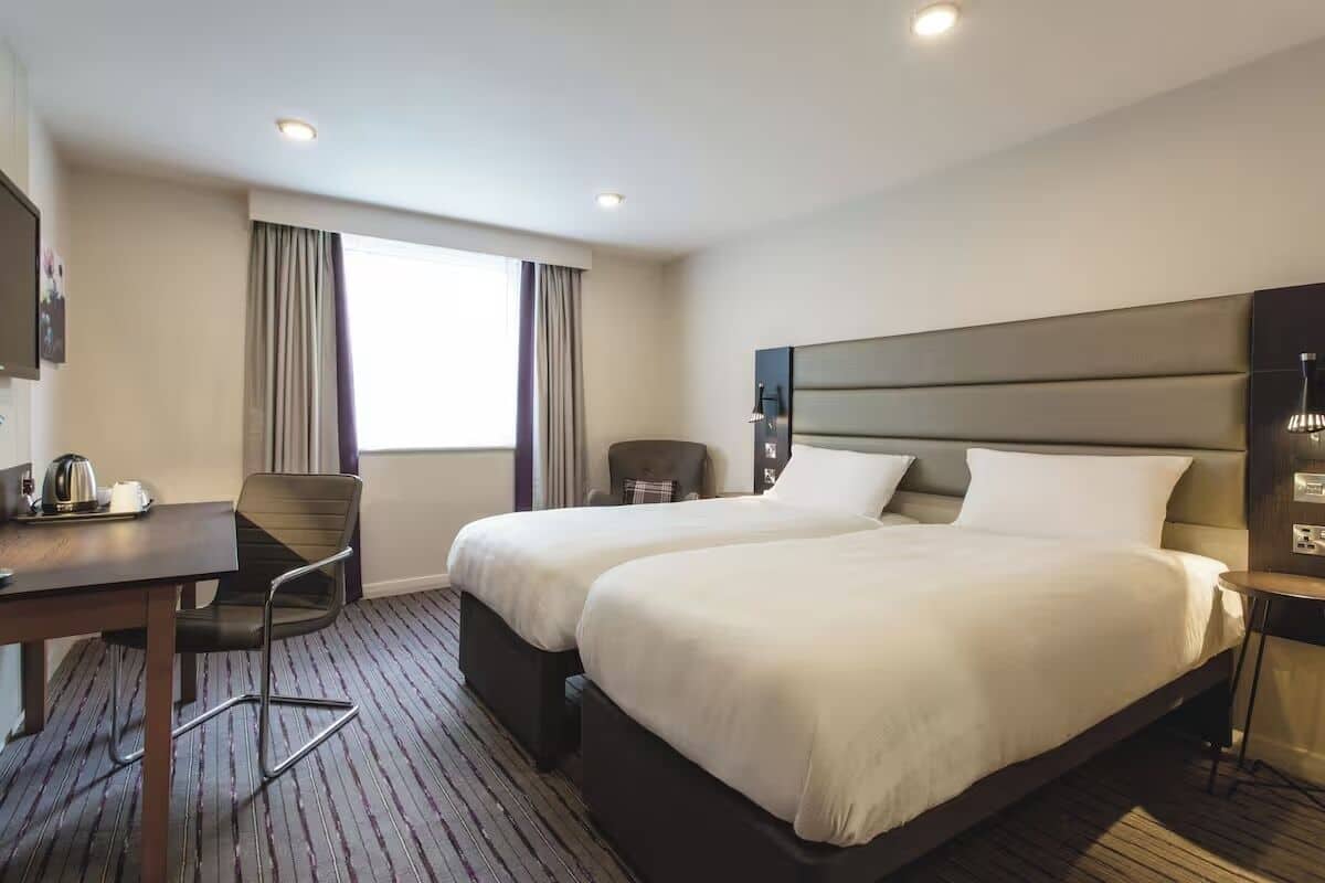 Ein Hotelzimmer mit zwei Betten, einem Schreibtisch mit Stuhl, einem kleinen Tisch mit Wasserkocher und einem Fenster mit Vorhängen.