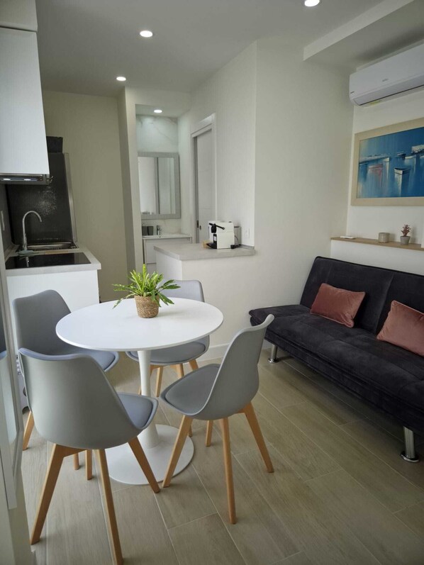 A compact living space with a kitchenette, a dining area with a round table and chairs, and a dark-colored sofa with cushions.