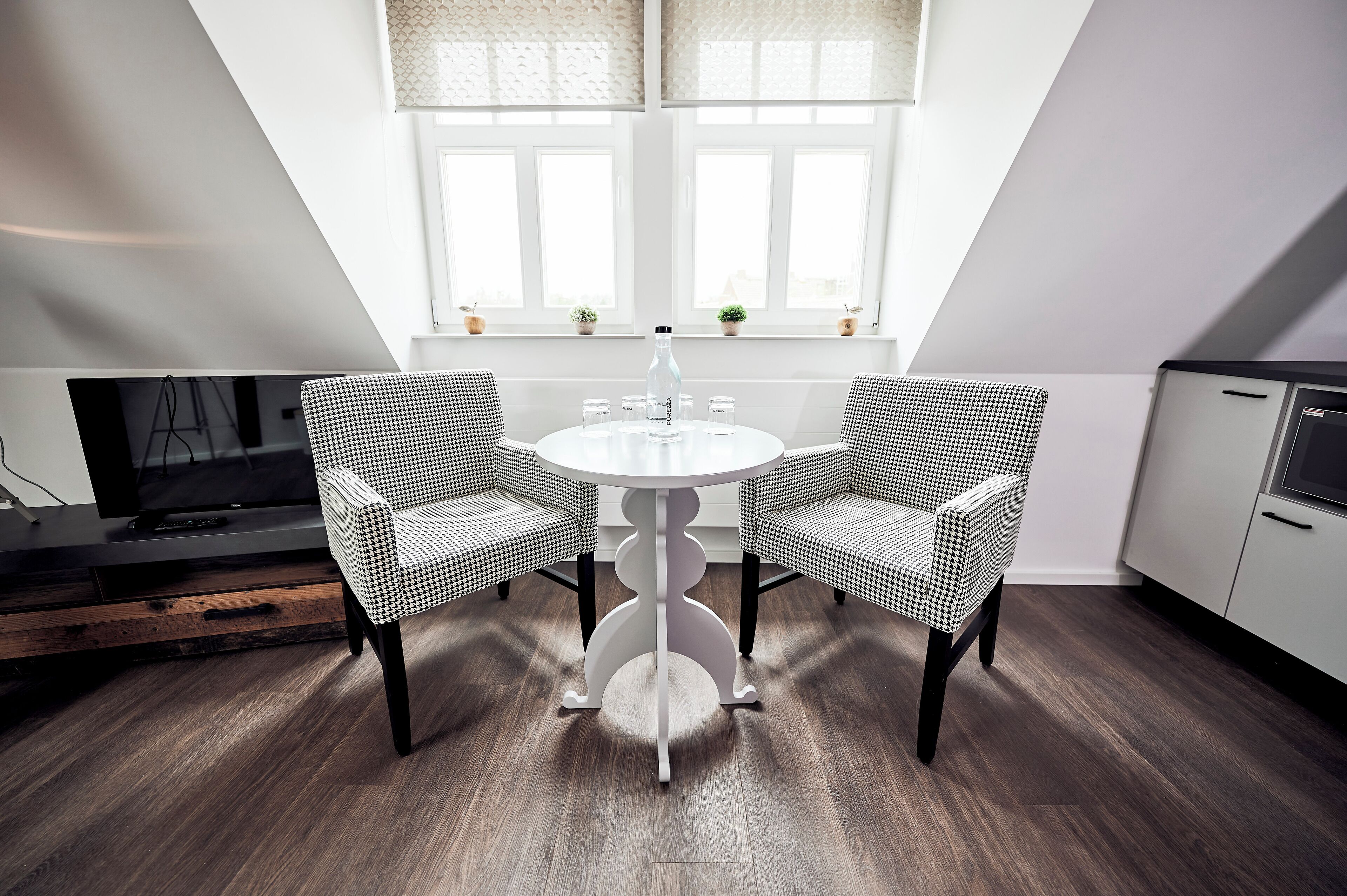 A dining area with a round table, two checkered chairs, a bottle, and a TV on a stand.