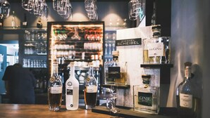 Uma área de bar com várias garrafas de bebidas alcoólicas, incluindo gin e uísque, e uma pessoa ao fundo.