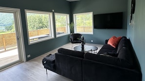 Ein modernes Wohnzimmer mit einer schwarzen Eckcouch, einem kleinen runden Tisch und einem flachbildfernseher, der an der Wand montiert ist. Große Fenster bieten einen Blick ins Freie.