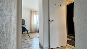 Un pasillo con piso de madera, una puerta blanca y vista a una habitación con cama y sillón.