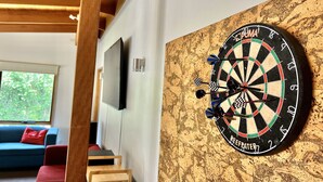 A dartboard mounted on a textured wall in a room with a wooden ceiling, a television, and a blue couch. - Albaski Lodge – Room 2 (Sawmill Settlement)