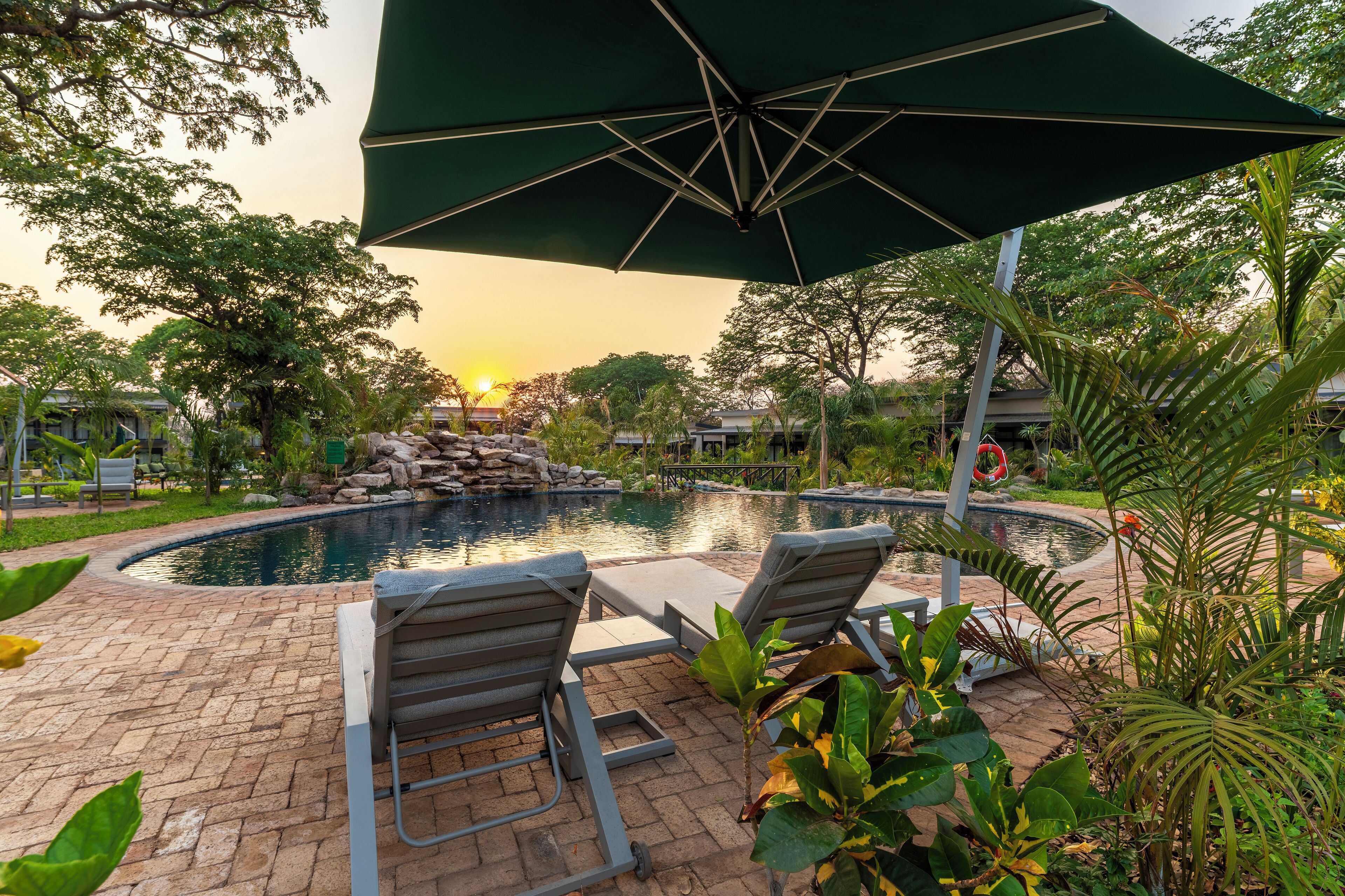 A pool area with lounge chairs and an umbrella, surrounded by greenery and a stone structure.