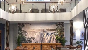 A spacious dining area with multiple wooden tables and chairs, a large waterfall mural on the wall, and modern pendant lighting. - Waterfalls Lodge (Victoria Falls)