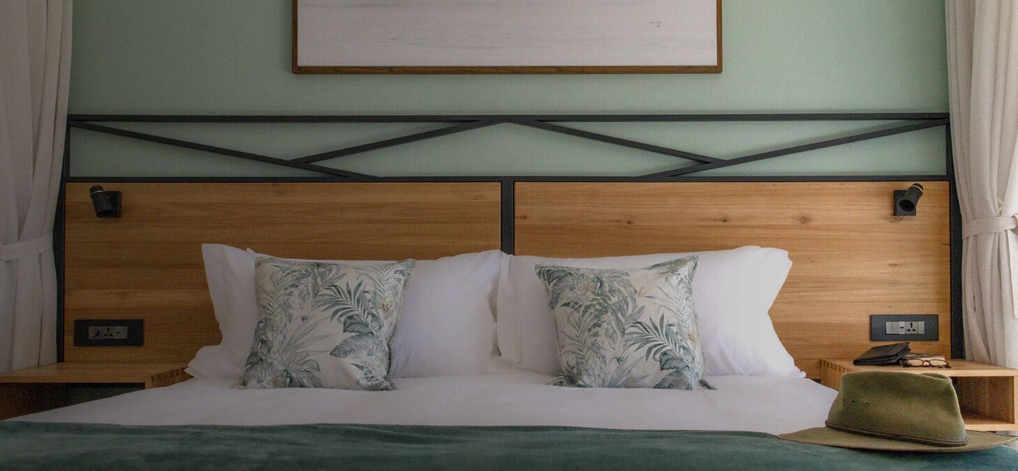 Una cama con cabecera de madera, dos almohadas blancas y una manta verde. Encima de la cama, hay un cuadro en la pared.