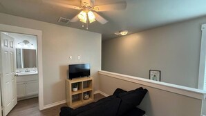 Ein Wohnzimmer mit schwarzem Sofa, einem Fernseher auf einem Holzgestell, einem Deckenventilator und einem Badezimmer mit Spiegel und Waschbecken.