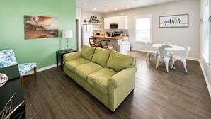 Un salon avec un canapé vert, un coin repas avec des chaises blanches, une cuisine avec des meubles blancs et un ventilateur de plafond.