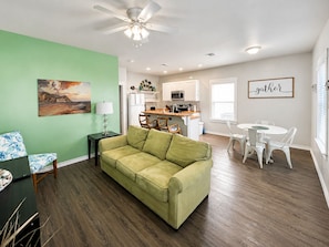 A living room with a green couch, a dining area with white chairs, a kitchen with white cabinets, and a ceiling fan.
