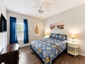 A bedroom with a bed, two bedside tables, a ceiling fan, a window with curtains, a wall-mounted TV, and a framed artwork.