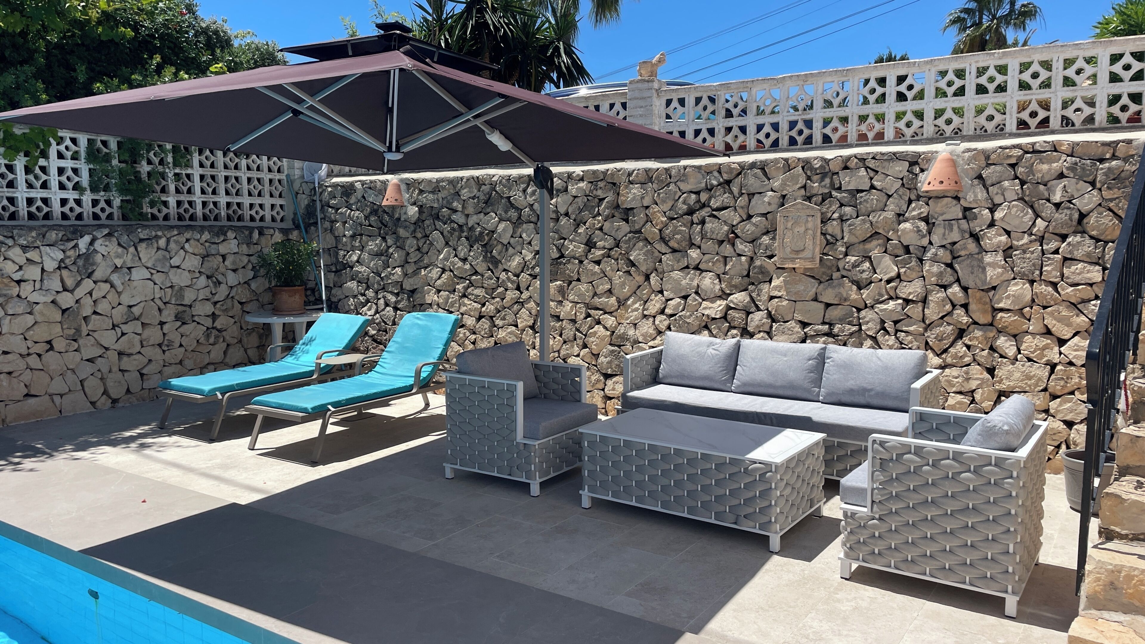 A patio area with a pool, stone wall, lounge chairs, a sofa, and a pergola.